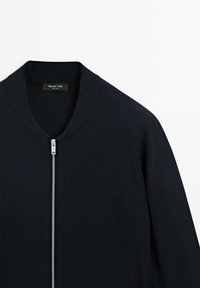Marinblå zip-up bomberjacka med ribbad krage, tillverkad av ett mjukt material, med en dragkedja i silverfärg och en minimalistisk design.