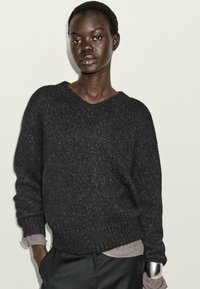 Schwarzer gestrickter Pullover mit V-Ausschnitt, lockerer Passform und strukturiertem Stoff. Das hellgraue Hemd darunter sorgt für einen Kontrast.