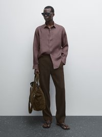 Braune Leinenbluse, lockere braune Hose, schwarze Sonnenbrille, trägt eine braune Tasche. Sandalen mit flachen Sohlen. Strukturierter Stoff, neutrale Töne.
