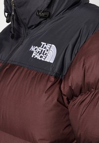 Doudoune bordeaux avec une partie supérieure noire, ornée d'un logo blanc. Le matériau est brillant et gonflé avec une texture matelassée.