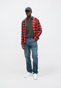 Červená a černá flanelová košile na kostky, šedé polo, modré džíny a bílé tenisky. Osoba má na hlavě šedou čepici a stojí před jednobarevným pozadím.