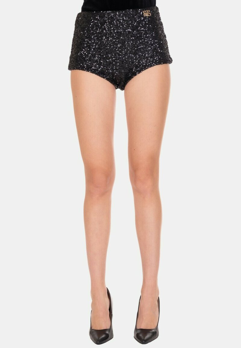 Shorts neri scintillanti a vita alta indossati con top nero e tacchi alti neri, mostrati su una modella in piedi contro uno sfondo chiaro uniforme.