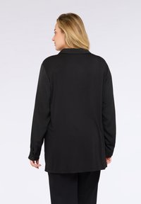 Donna con capelli biondi che indossa una maglietta nera a maniche lunghe e pantaloni neri, di spalle contro uno sfondo chiaro e semplice.