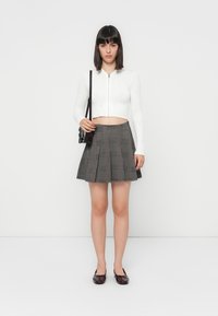 Hvit kortet genser med glidelås, grå rutete plissert skjørt og mørke loafers. Modellen holder en liten svart veske. Minimalistisk bakgrunn.