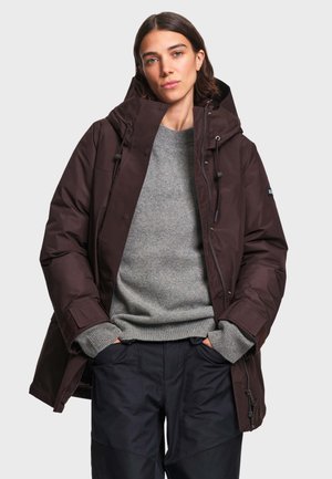 Femme portant une veste d'hiver marron foncé à capuche par-dessus un pull gris et un pantalon noir, debout les mains dans les poches devant un fond uni.