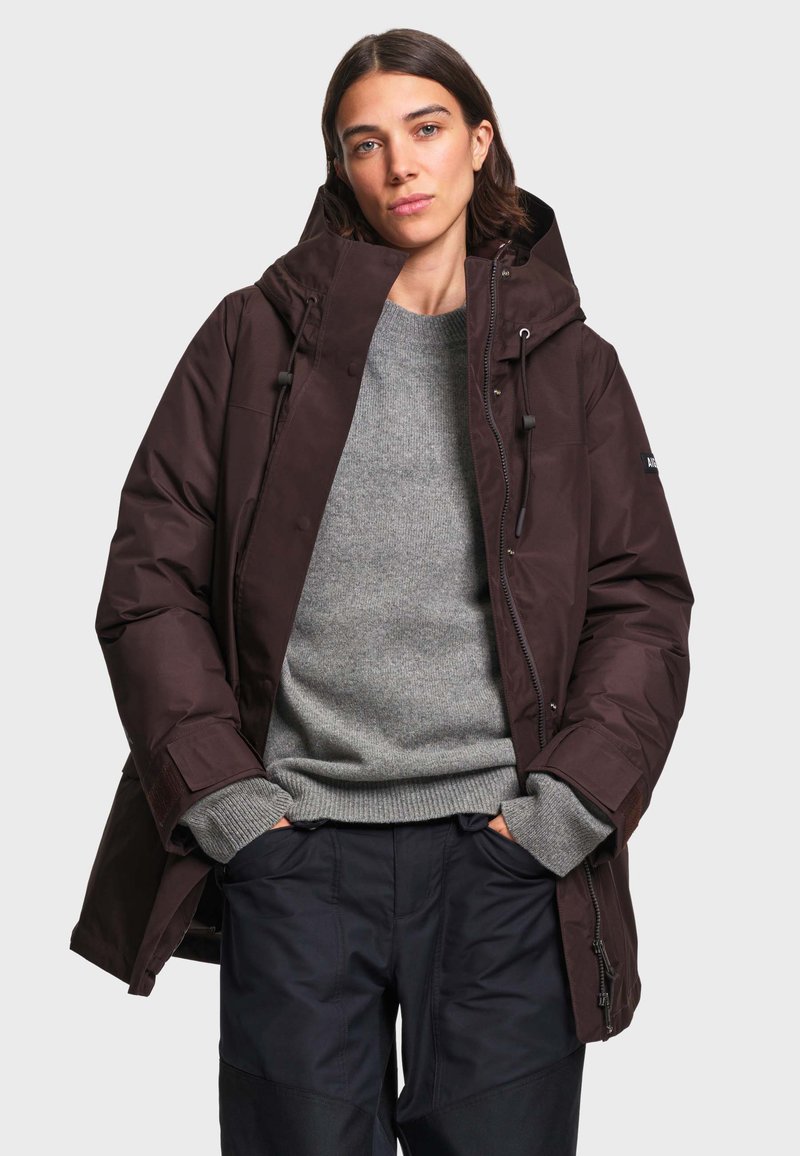 Femme portant une veste d'hiver marron foncé à capuche par-dessus un pull gris et un pantalon noir, debout les mains dans les poches devant un fond uni.