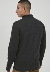 Suéter de color gris oscuro con un cuello alto, puños y dobladillo acanalados, y una tela suave y texturizada. Pequeño logo en la manga izquierda.