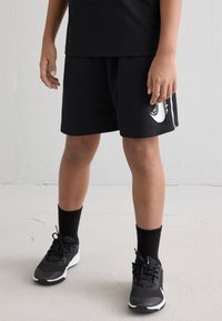 Pantalones cortos deportivos negros con un logo blanco, combinados con calcetines negros y zapatillas grises. Los pantalones cortos tienen un corte relajado y una textura suave.