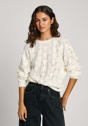 Cremefarvet strikket sweater med et blonderet mønster og ribbestrikkede manchetter, parret med højtsiddende sorte jeans med nitter på lommerne.