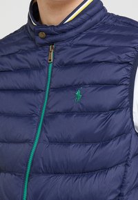 Námořnická puffer vesta s vertikálním zeleným zipem. Má žebrovaný límec s žlutými a bílými pruhy a zelené vyšití loga na hrudi.