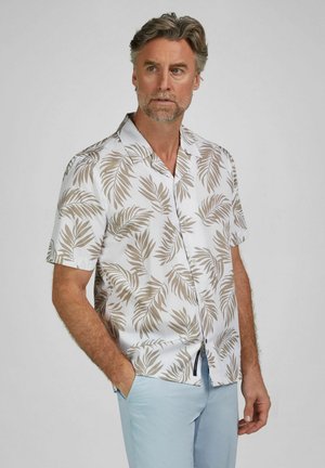 Man draagt een wit shirt met korte mouwen en beige bladpatroon en lichtblauwe broek, staat met één hand in zijn zak en kijkt zijwaarts.