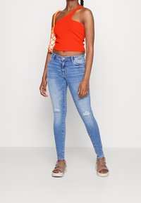 Červený crop top na jedno rameno, světle modré roztrhané skinny džíny a hnědé sandály s páskem kolem kotníku. Oranžová síťová taška dodává kontrast.