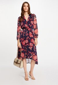 Robe imprimée florale en bleu marine avec des accents rouges, dotée d'un décolleté en V, de longues manches transparentes et d'une taille cintrée. Associée à un sac à main doré.