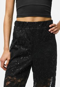 Crne čipkaste hlače s cvjetnim uzorkom, ukrašene šljokicama i elastičnim pojasom. Prozirna tekstura pojačava vizualni dojam.