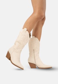 Białe kowbojskie buty z wyszywanymi kwiatowymi wzorami, spiczastym noskiem i bocznym zamkiem błyskawicznym. Charakteryzują się brązowym, stylizowanym obcasem.