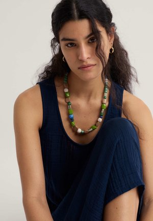 Colier cu mărgele multicolore, cu nuanțe de verde, albastru și tonuri neutre, cu forme și dimensiuni variate, pe un fundal de țesătură texturată albastru închis.