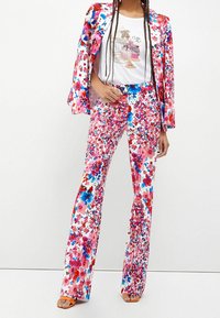 Vrouw in een heldere bloemenpak met bijpassende blazer en broek, witte grafische t-shirt en oranje open teen hakken tegen een witte achtergrond.