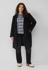 Frau mit lockigem Haar, die einen schwarzen Mantel, ein blau kariertes Hemd, schwarze Jeans und weiße Turnschuhe trägt, steht vor einem schlichten hellen Hintergrund.