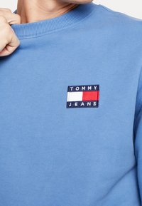 Persona che indossa una felpa leggera blu di Tommy Jeans, mentre tiene il collo della felpa con una mano, mostrando il logo del marchio sul petto.