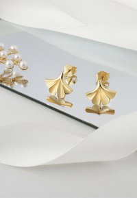 Guldfärgade örhängen formade som ginkgoblad med en slät, blank yta och fiskkroksspärr. Placeras på en reflekterande yta.