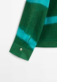 Poignet de chemise à manches longues en tissu tie-dye vert et bleu sarcelle avec un seul bouton beige sur le poignet.