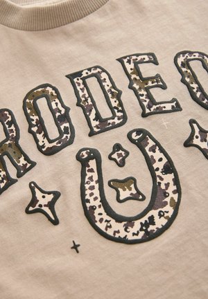 T-shirt in cotone beige con scritte scure "RODEO", caratterizzato da un design a forma di ferro di cavallo testurizzato con stampa animale e da forme astratte in marrone e beige.