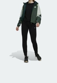 Vihreä ja valkoinen hupullinen takki, musta paita, mustat kapealahkeiset housut ja mustat urheilukengät. Takissa ja housuissa on logo.