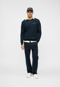 Sudadera navy con logo, superpuesta a una camisa de cuello amarilla clara, combinada con pantalones oscuros y zapatillas blancas. Una gorra gris completa el look.
