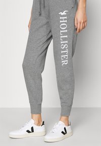 Šedé tepláky s bílým logem "Hollister" na boku, s manžetami na kotnících; v kombinaci s bílými teniskami s černými akcenty.