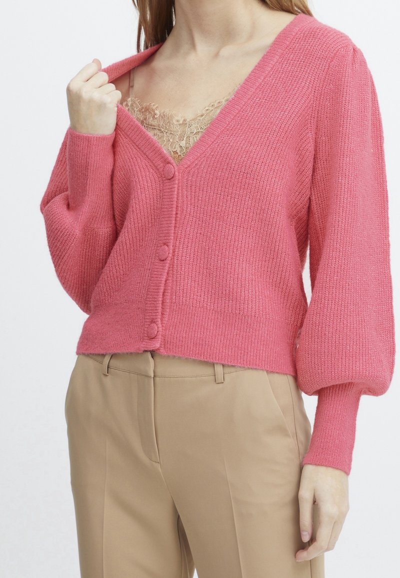 Femme portant un cardigan rose boutonné sur un haut beige orné de dentelle et un pantalon beige taille haute, tenant le col du cardigan d'une main.