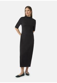 Robe noire, au genou, avec un col montant et des manches courtes. Texture lisse, silhouette ajustée, associée à des ballerines noires.