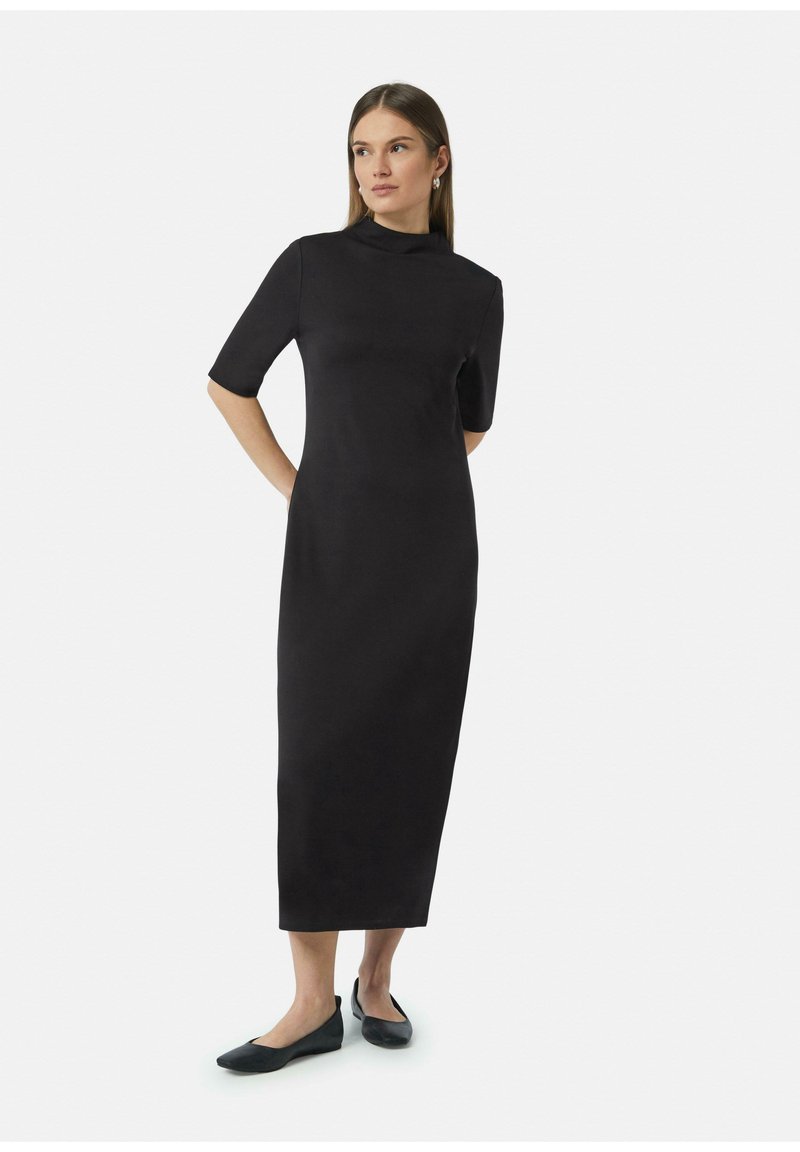 Robe noire, au genou, avec un col montant et des manches courtes. Texture lisse, silhouette ajustée, associée à des ballerines noires.