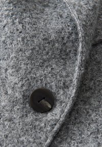 Nahaufnahme eines grauen strukturierten Stoffes mit einem großen schwarzen Knopf. Das Material hat ein fusseliges Aussehen mit sichtbaren Schlaufen und einer weichen Oberfläche.