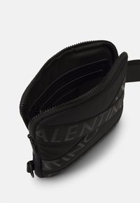 Svart dragkedjeväska med texturerat material, flera fack och en framträdande logotypdesign. Justerbar rem ingår.