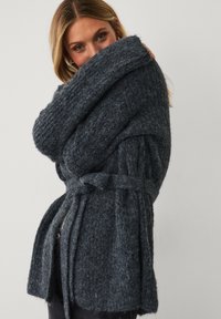 Frau mit blonden Haaren, die eine dunkelgraue, grob gestrickte Strickjacke mit einem Gürtel trägt und teilweise ihr Gesicht mit dem Ärmel vor einem schlichten Hintergrund verdeckt.