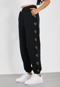 Person i svarta joggers med vita hjärtlinjer längs sidan, en vit kort topp och vita sneakers, stående på grått golv.