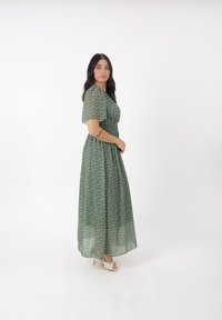 Maxikleid aus transparentem grünen Stoff mit floralen Mustern, kurzen Puffärmeln und figurbetontem Taillenbereich. Kombiniert mit eleganten, hellen Absatzschuhen.
