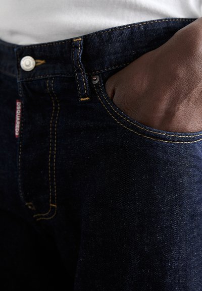 Dunkelblaue Denim-Jeans mit kontrastierenden gelben Nähten, einer Fronttasche und einem metallischen Verschluss. Beinhaltet ein Markenlabel an der Seite.