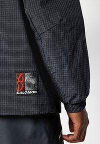 Jachetă neagră tip windbreaker cu model în grilaj, manșete elastice și design ajustat. Prezintă un patch cu logo negru și roșu pe partea stângă.