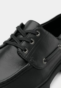 Μαύρο δερμάτινο παπούτσι με λεία υφή, σχέδιο δέσιμου με κορδόνια, ασημένιες οπές και αντίθετη ραφή κατά μήκος της σόλας.