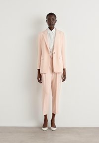 Completo su misura rosa chiaro con giacca monopetto e pantaloni slim-fit, abbinato a una camicia bianca e tacchi bianchi. Tessuto con una texture liscia.