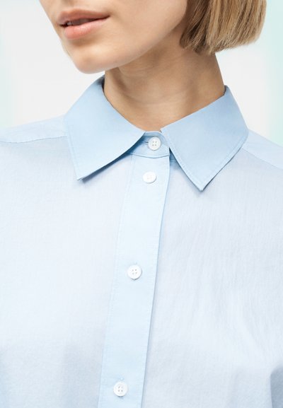 Κοντινό πλάνο ενός ατόμου που φοράει ανοιχτό μπλε πουκάμισο με κουμπιά και κλασικό γιακά, εμφανίζοντας τον λαιμό και το κάτω μέρος του προσώπου.