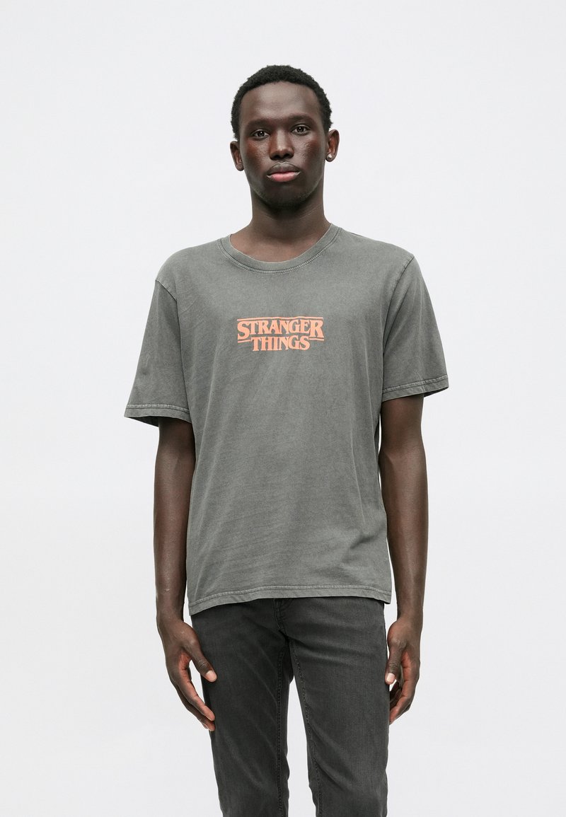 Gray cotton t-shirt with short sleeves, featuring an orange "STRANGER THINGS" logo centered on the chest. Simple, relaxed fit design.