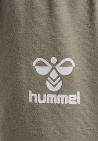 Olivgrön tyg med en strukturerad yta som har en framträdande vit logotyp med två vingar och en bladdesign, med texten "hummel" nedanför.