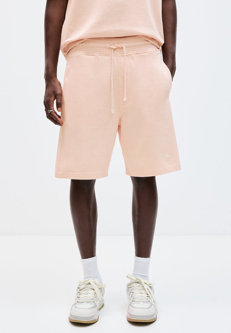 Pantaloni scurți dintr-un amestec de bumbac de culoare piersică, cu un tiv elastic, șnur, buzunare laterale și un mic detaliu cu logo. Asortați cu adidași și șosete albe.