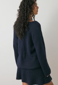 Tumedat sinise värvi kudumine-sweater, millel on lõdvestatud lõige ja tekstureeritud kangas, kellel on pikad varrukad ja ümmargune kaelus. Kantud sobiva seeliku kõrval.