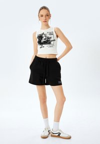 Top corto blanco con diseño gráfico, combinado con pantalones cortos negros con cordón. El modelo lleva calcetines blancos y zapatillas informales.