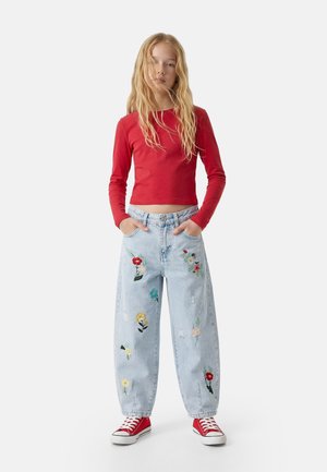 Junge Person mit langen blonden Haaren trägt ein rotes langärmeliges Crop-Top, hellblaue Jeans mit Blumenstickereien und rote Sneakers, steht mit den Händen in den Taschen.