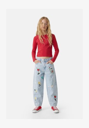 Joven con cabello rubio largo lleva un crop top rojo de manga larga, vaqueros azul claro con bordados florales y zapatillas rojas, de pie con las manos en los bolsillos.