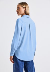 Camicia azzurra chiaro con maniche lunghe, colletto e dettaglio della piega posteriore, realizzata in tessuto liscio, abbinata a pantaloni in denim blu.